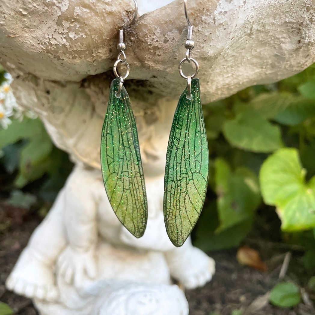 Sparkly glitter green dragonfly wing earrings hanging from a garden ornament.