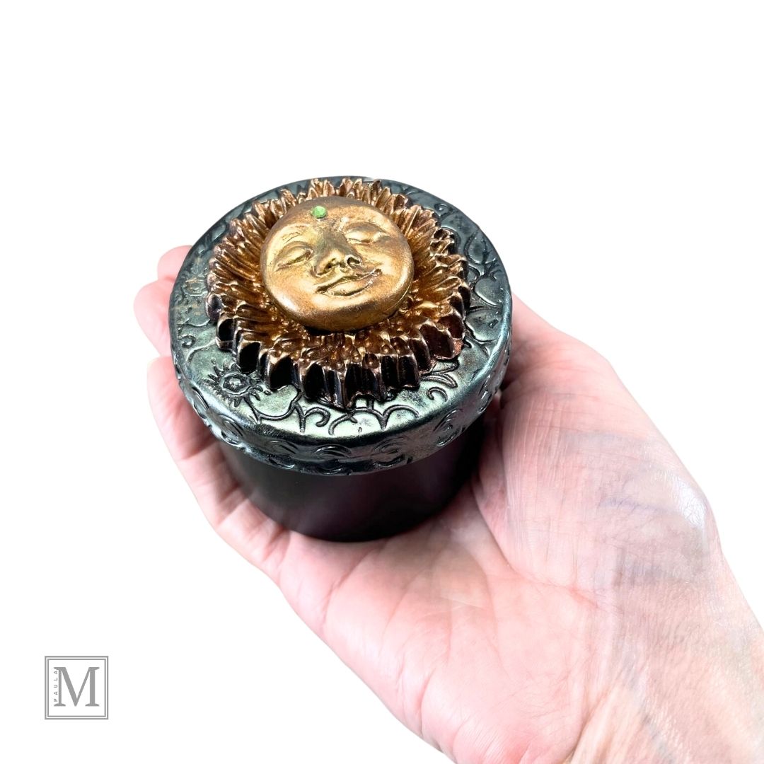 Black storage tin with decorative polymer clay lid with gold sun face design held in model's hand for size reference
