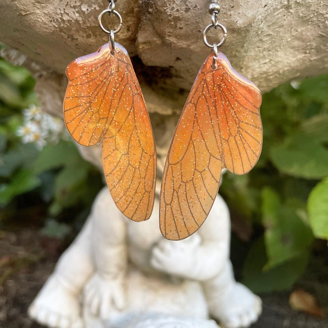 orange ombre sparkly glitter plastic fairy wing earrings hanging from a fairy garden ornament