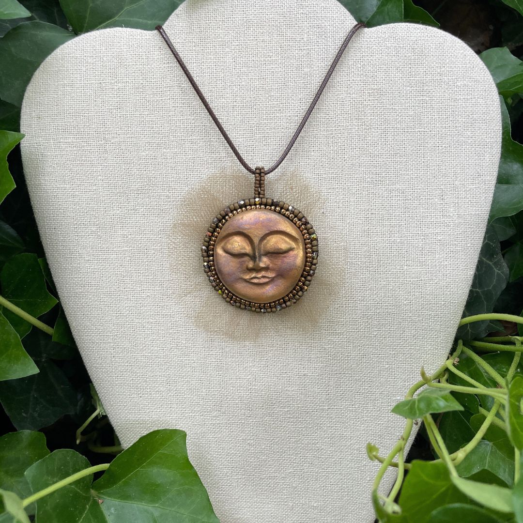 Beaded sun goddess statement pendant with sleeping face and gold beading and tulle around the edge . On a display stand with greenery.