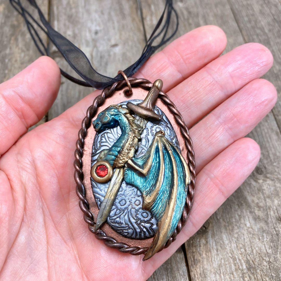 Handmade fantasy dragon and sword pendant necklace in teal, bronze, and red tones. High fantasy jewelry gift for dragon lovers and cosplay fans. Held in a hand.