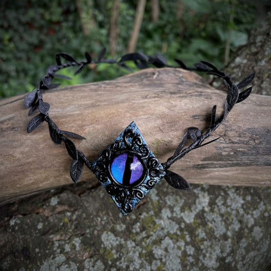 Purple dragon on a diamond shape textured clay base on a black leaf wire headband resting on a tree branch.