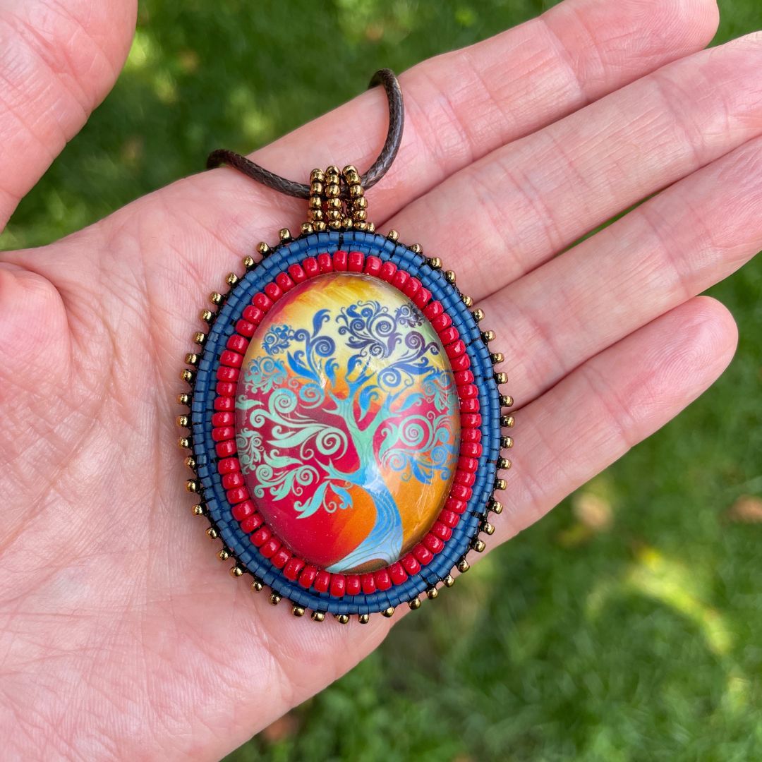 Ovsl pendant with swirly whimsical blue tree with gold and red sunset with blue, red and black glass seebead edging held in a hand.