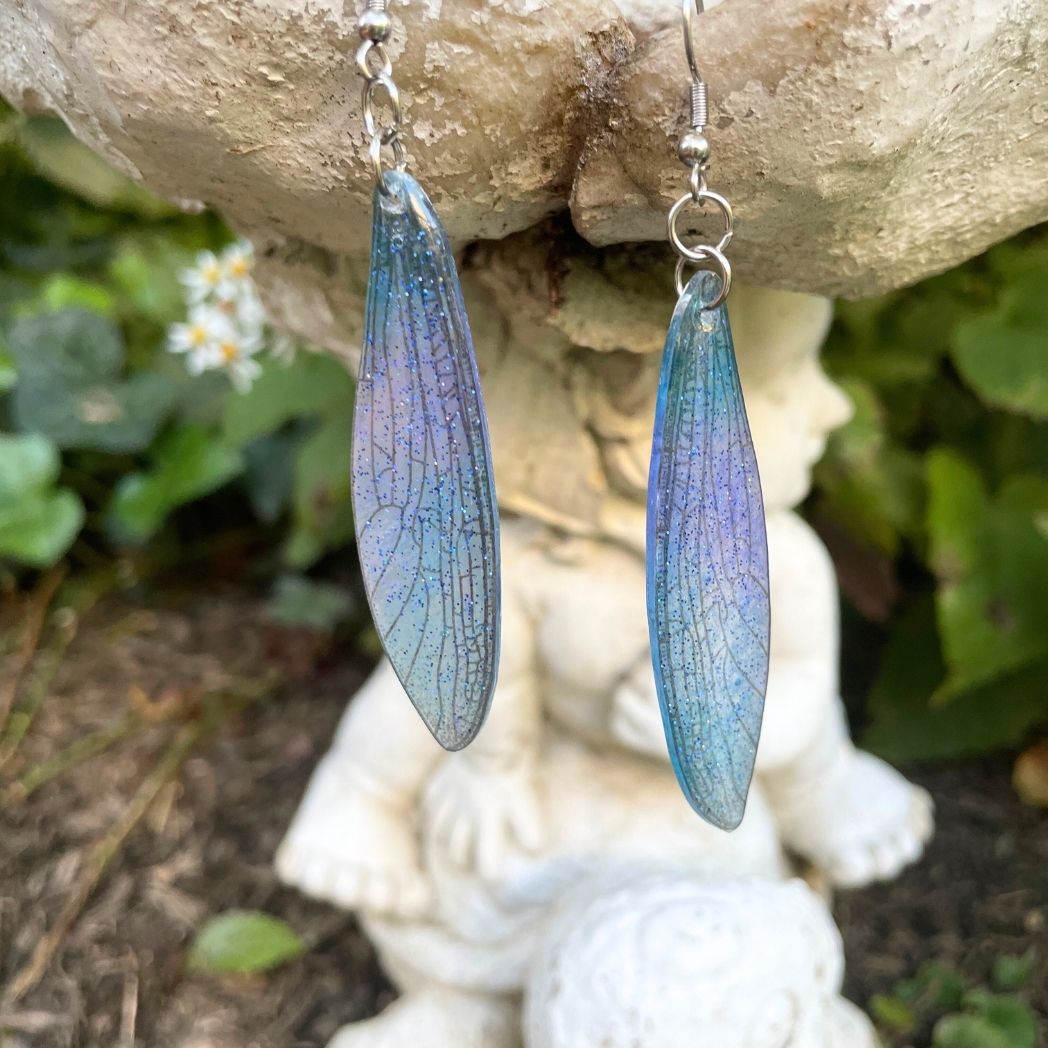 Sparkly glitter turquoise and pink dragonfly wing earrings hanging from a garden ornament.