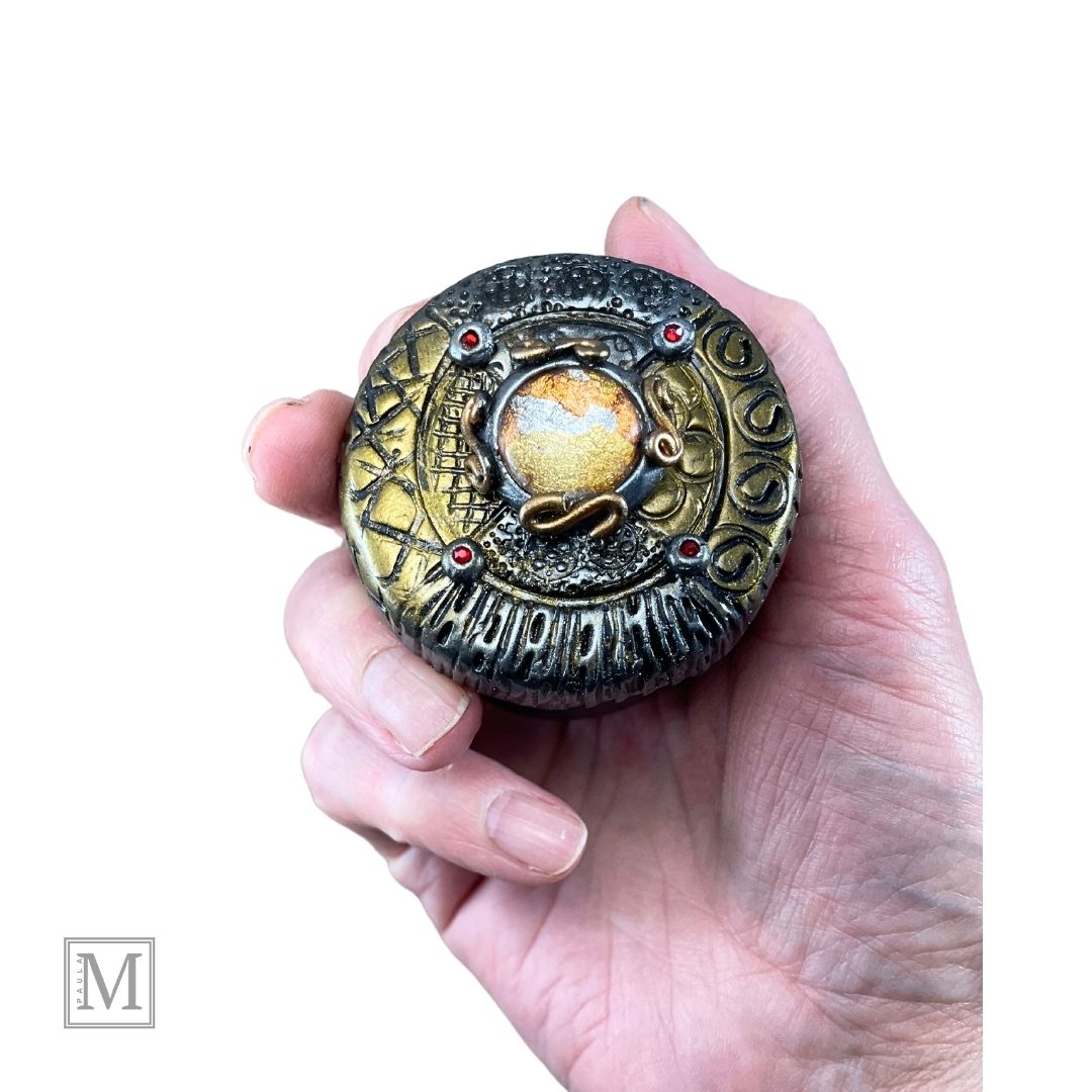 2oz black aluminum storage tin with handcrafted polymer clay lid with foiled glass center orb and gold and silver stamped details and red crystal accents.  In hand for size reference.