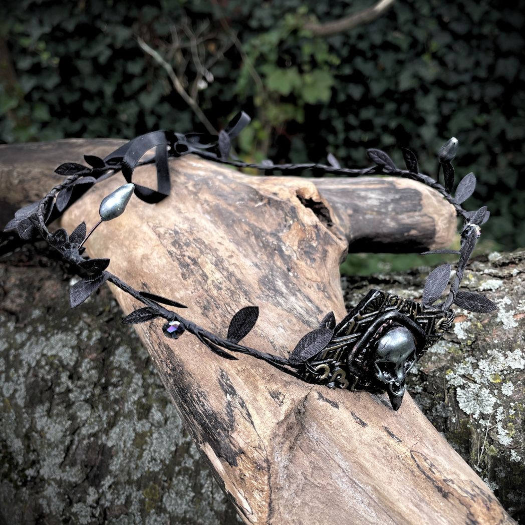 A silver raven skull on textured gold highlighted diamond shape focal on a black leaf circlet with black crystals and silver flower buds. Circlet is resting on a tree branch.