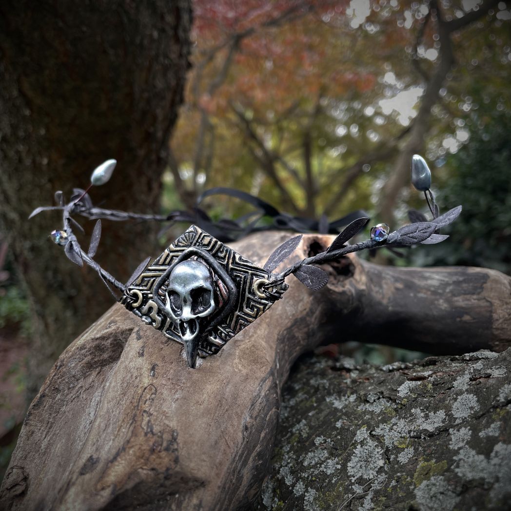 A silver raven skull on textured gold highlighted diamond shape focal on a black leaf circlet with black crystals and silver flower buds. Circlet is resting on a tree branch.