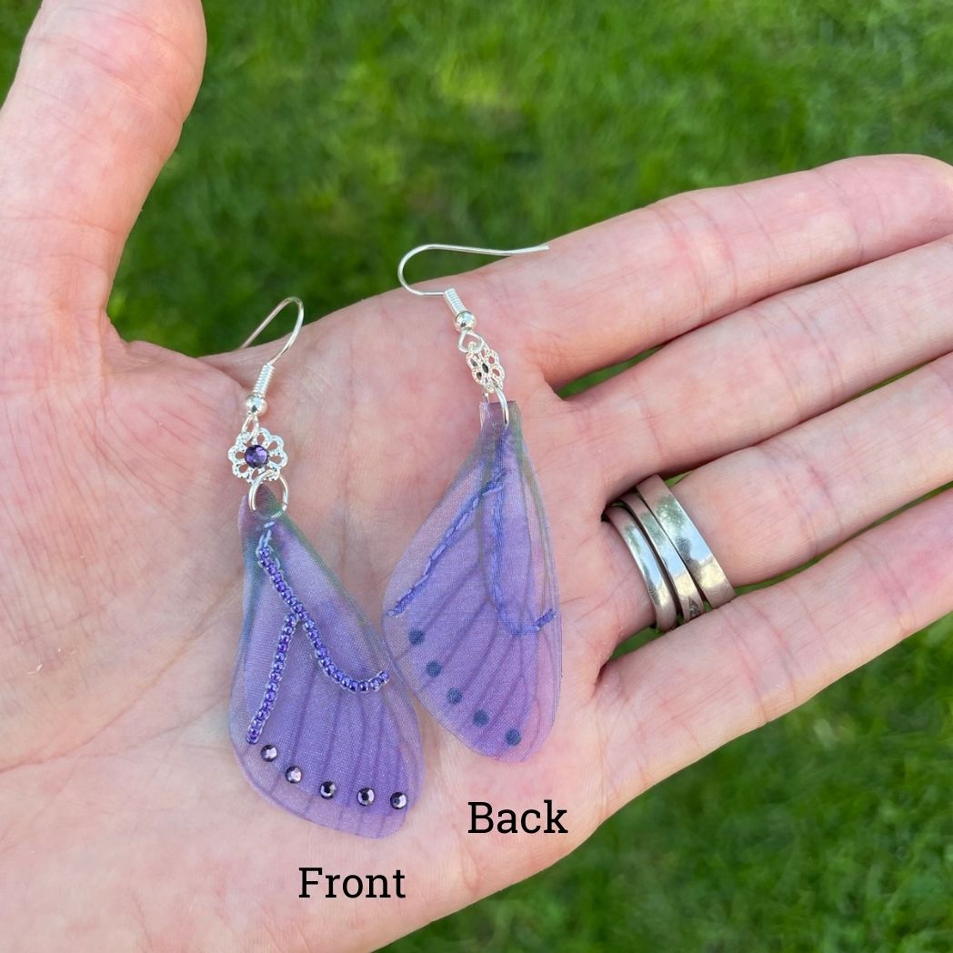 purple beaded fabric wing earrings with purple crystals and a flower bead with purple crystal above wings connecting to the ear wires held in a hand