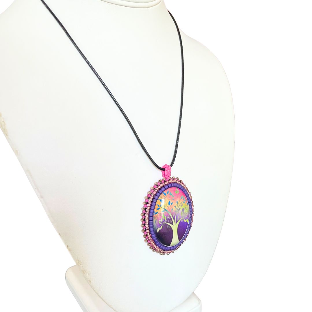 Oval pendant with whimsical tree with purple, red and pink background with pink and purple glass seed bead edging on a black cord necklace.