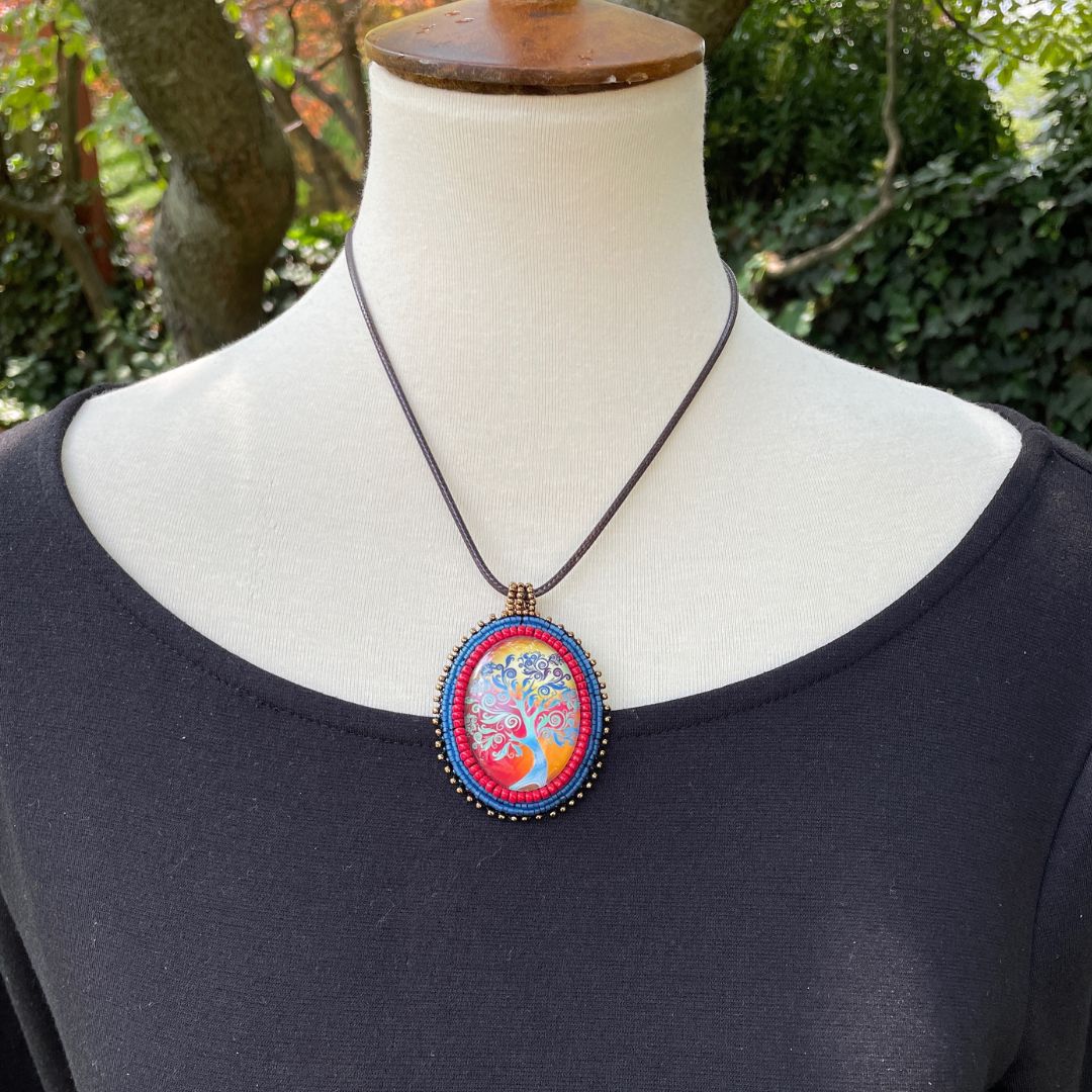 Ovsl pendant with swirly whimsical blue tree with gold and red sunset with blue, red and black glass seebead edging on a mannequin with black dress