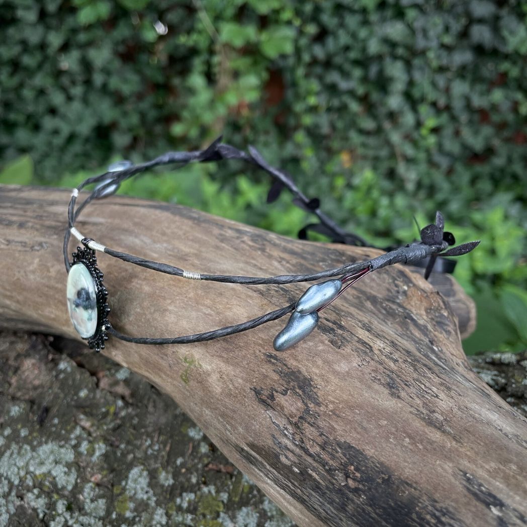Black wire crown with oval cabochon focal of a raven and skull resting on a tree branch.