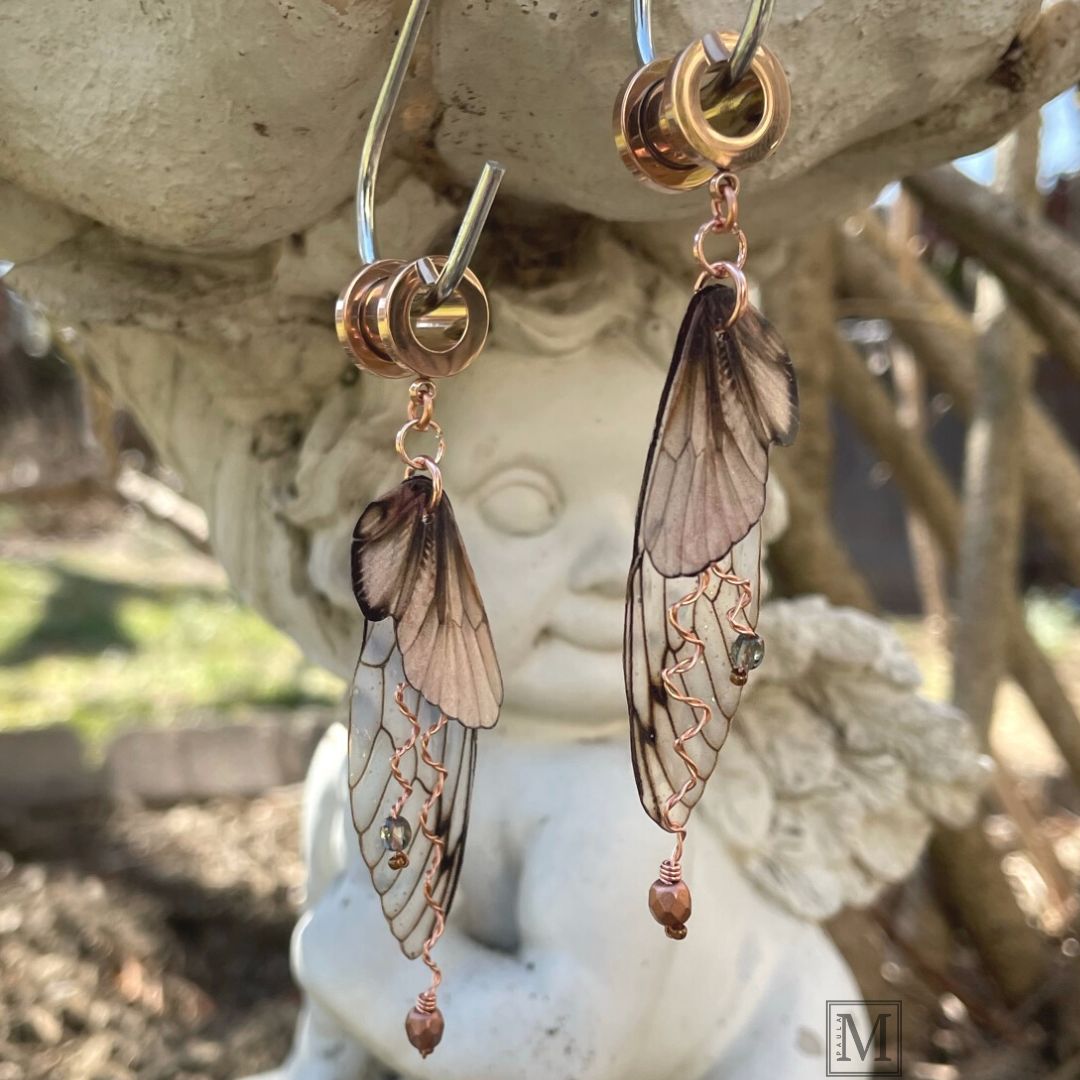 Double fairy wing earrings in brown with copper accents, with ear tunnels for stretched ears. Earrings are hanging from a garden angel ornament.