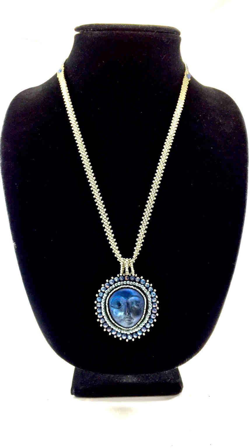 A necklace with a blue moon face surrounded by silver and blue beadwork on a long silver seed bead chain on a black jewelry bust.