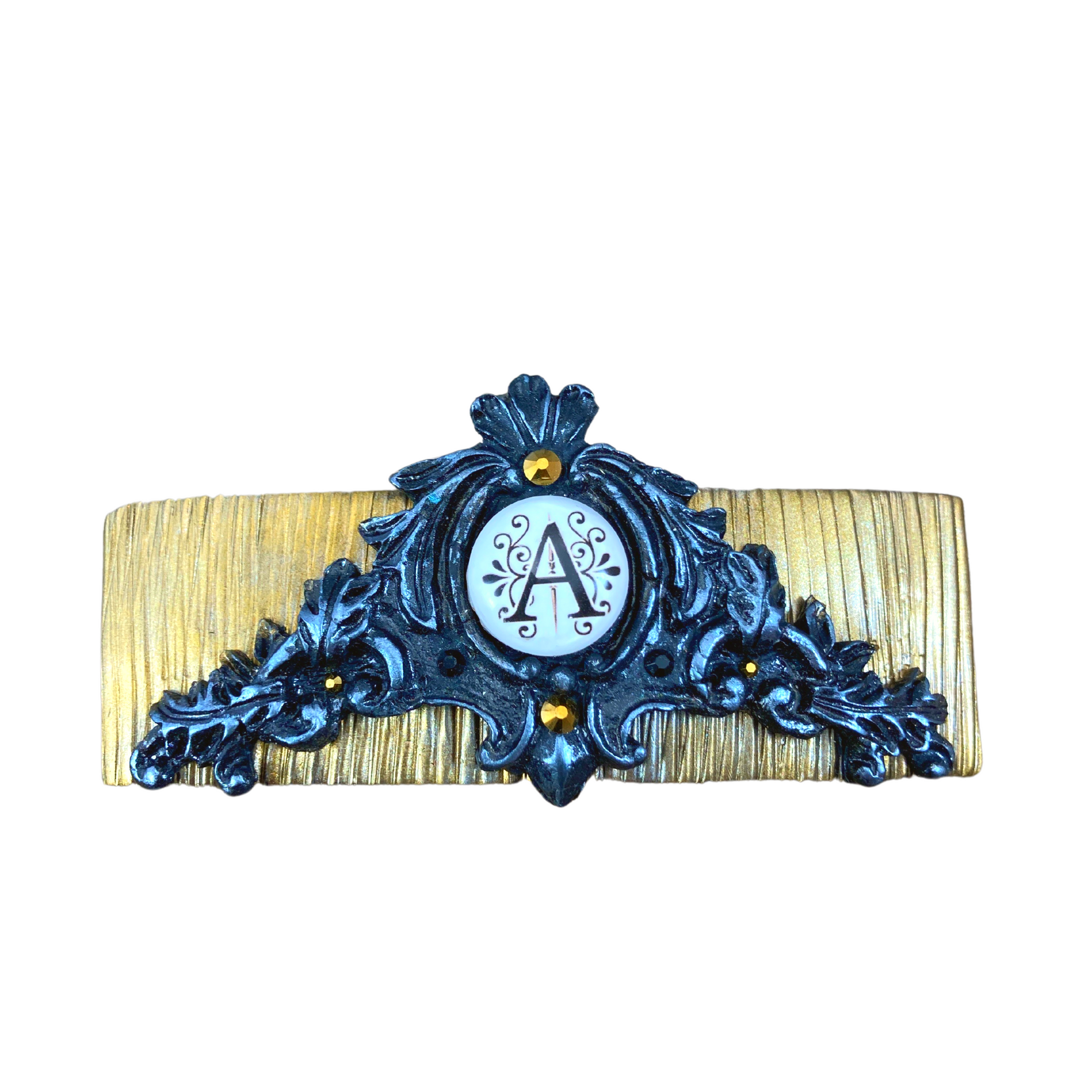 A gold clay base hair clip with a filigree design and black and silver in the center a graphic with a letter A.