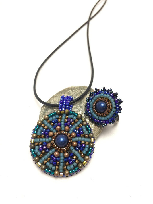 Seed beaded mandala pendant necklace and ring set in blue and green resting on a pebble.