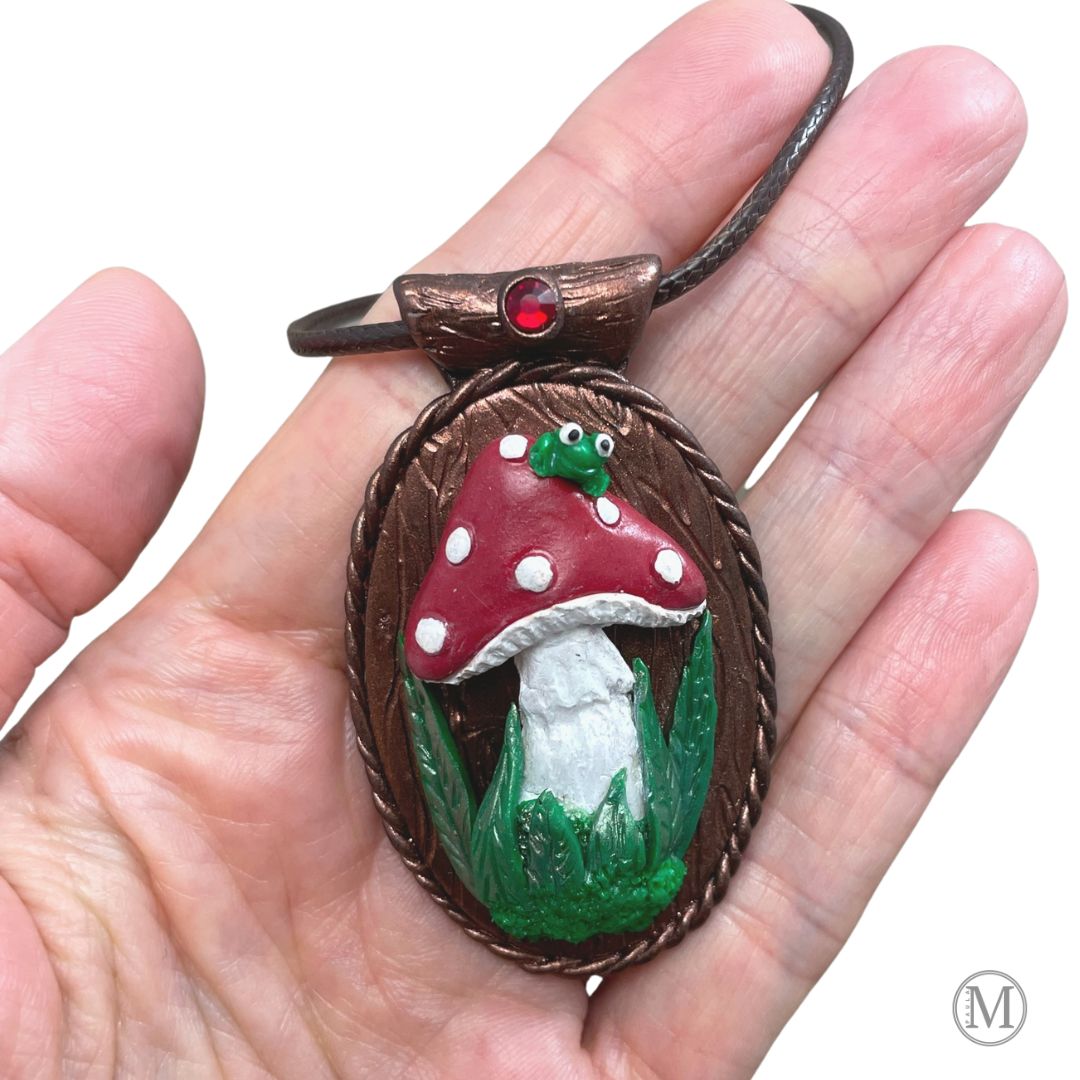Oval pendant with red spotted mushroom with grass and moss at stem with little green frog sitting on top of the mushroom. Brown cord necklace through a tube bail with a red crystal held in a hand.