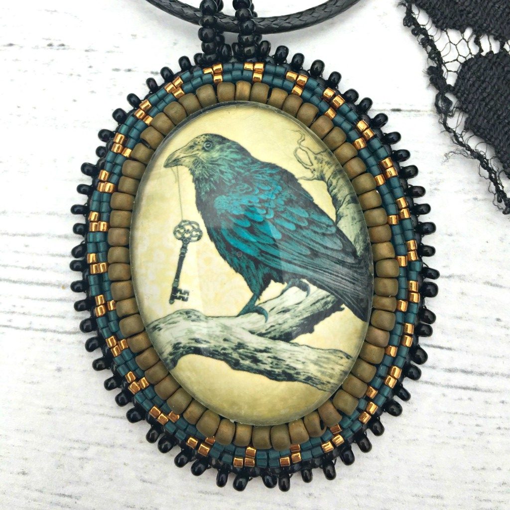 Necklace - Antiqued oval image of blue raven sitting on branch with a key in his beak surrounded by matte gold, blue and black beading. A piece of black lace is next to the pendant.