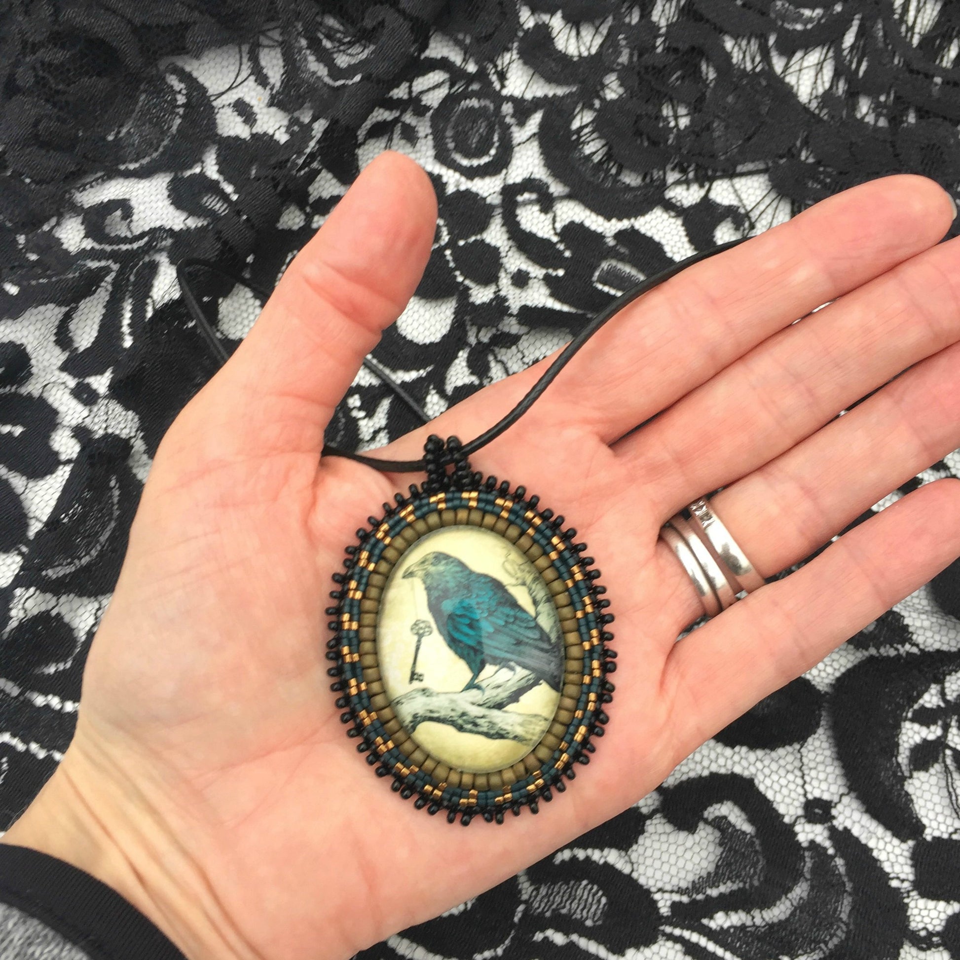 Necklace - Antiqued oval image of blue raven sitting on branch with a key in his beak surrounded by matte gold, blue and black beading. Necklace is held in a hand for size reference. A piece of black lace is in the background 