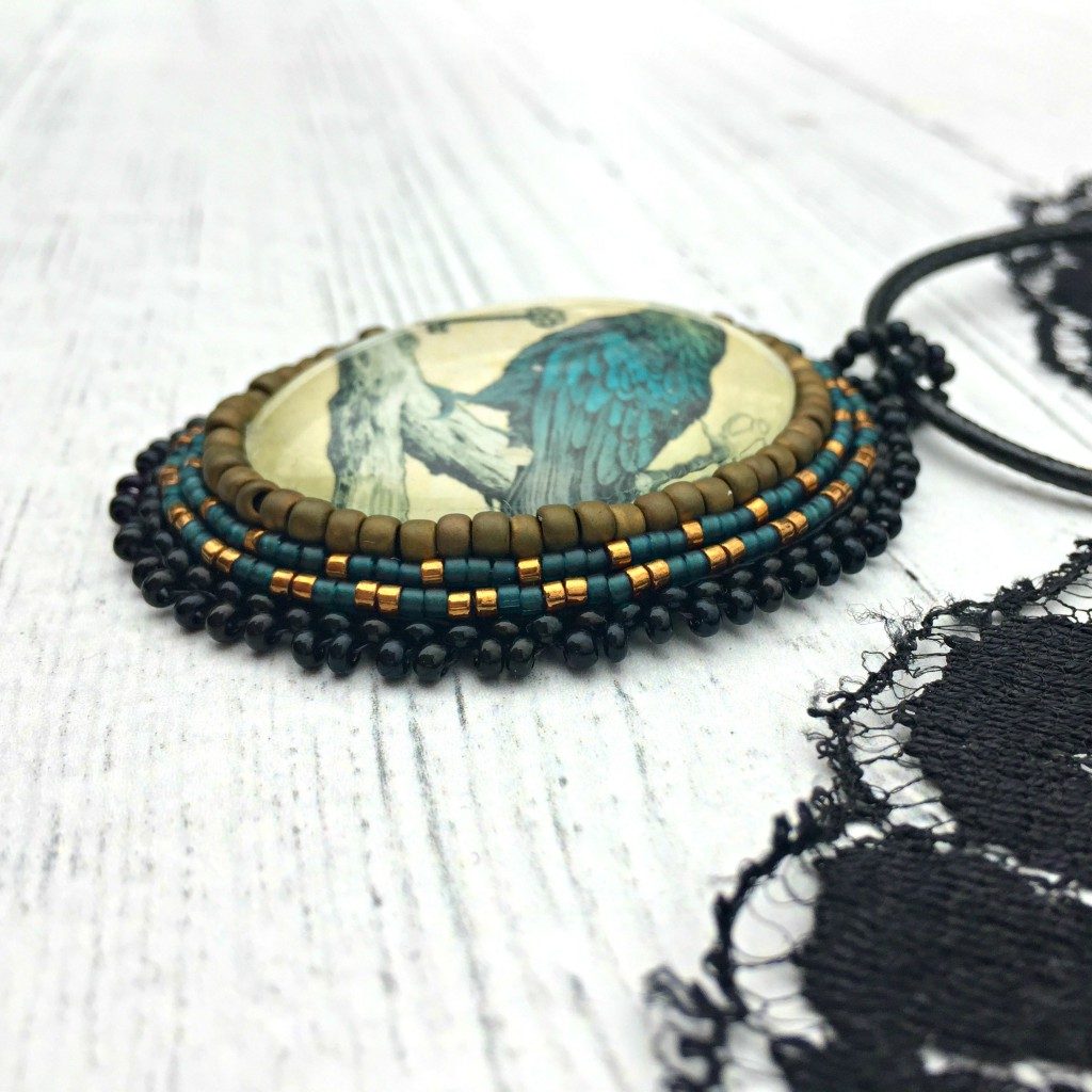 Side profile of necklace - Antiqued oval image of blue raven sitting on branch with a key in his beak surrounded by matte gold, blue and black beading. A piece of black lace is next to the pendant.