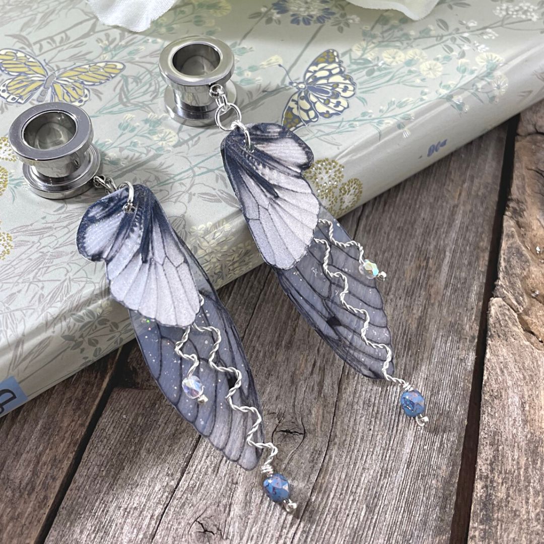 Double fairy wing earrings with silver accents, with ear tunnels for stretched ears. Earrings are leaning against a book.