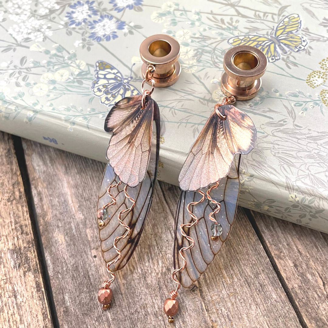 Double fairy wing earrings in brown with copper accents, with ear tunnels for stretched ears. Earrings are leaning against a book.