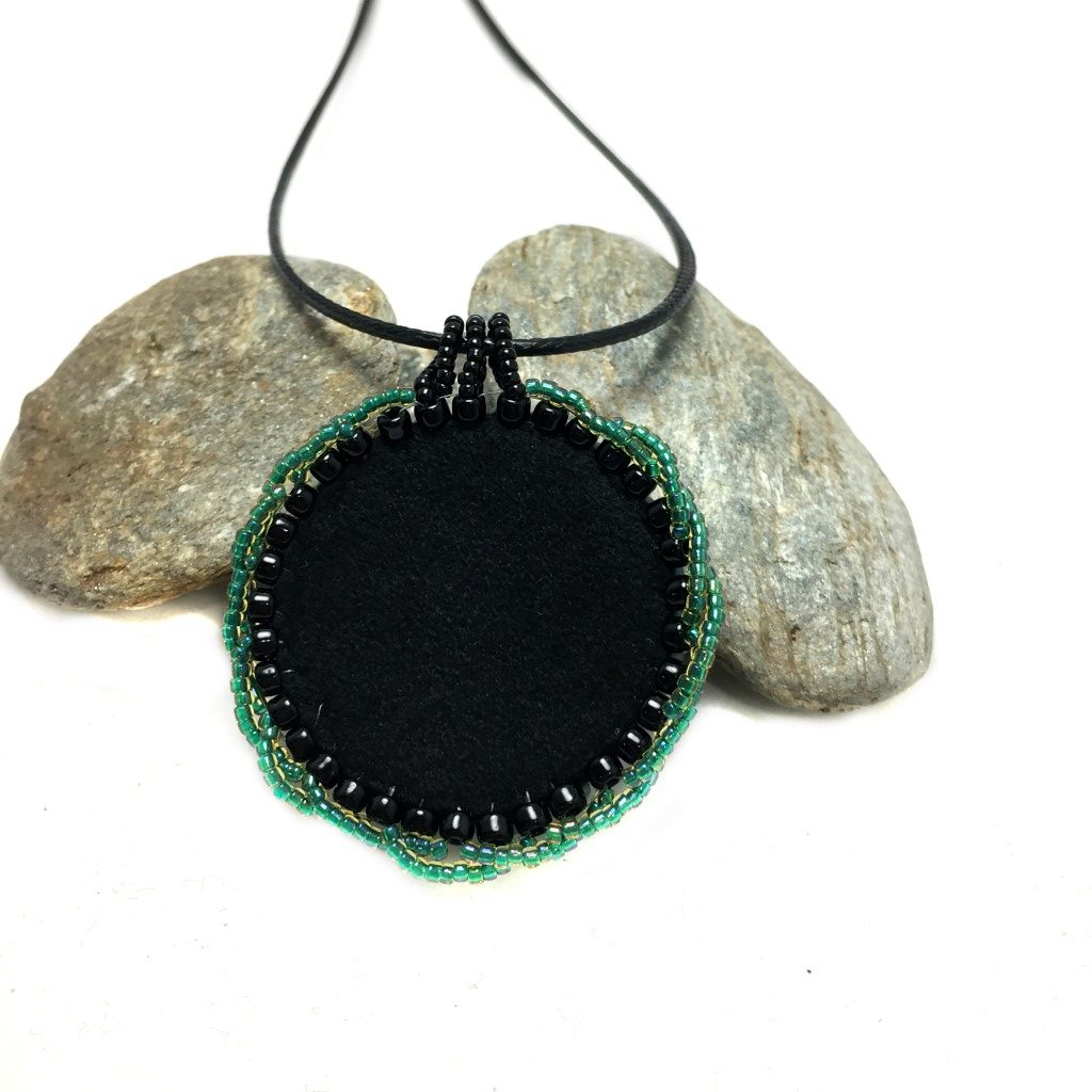 Back of Celtic pendant, showing black, faux, suede, and green, seeded twisted edging, resting onto pebbles.