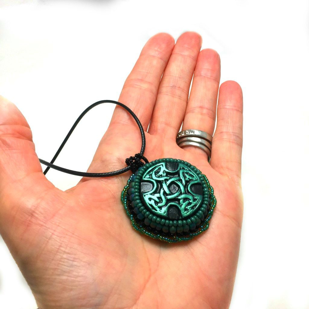 Green and black Celtic cross pendant with green twisted beaded edging held a hand for size reference.