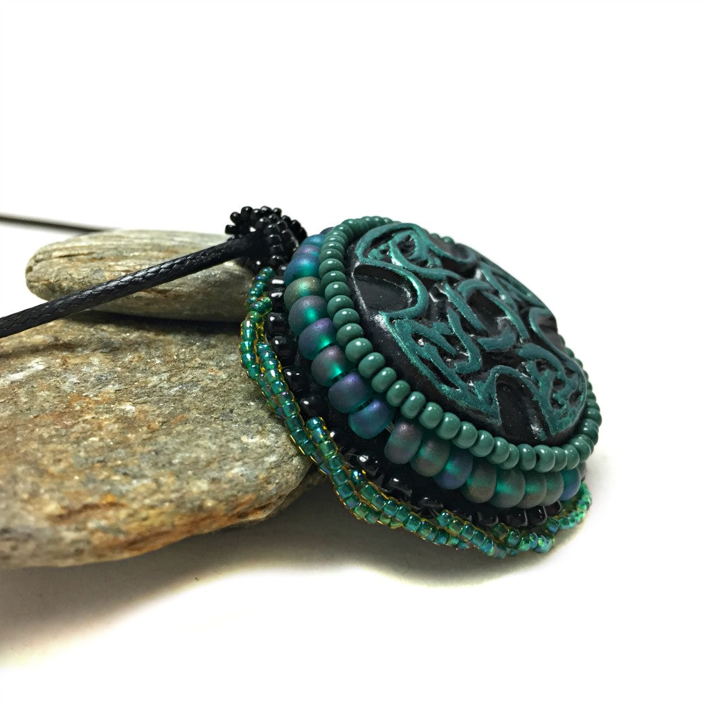 Side profile of Green and black Celtic cross pendant with green twisted beaded edging resting on a pebble.