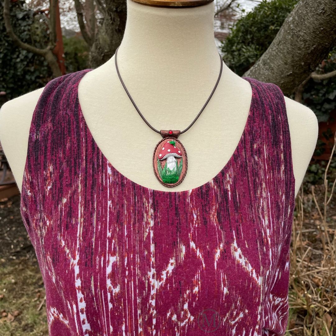 Oval pendant with red spotted mushroom with grass and moss at stem with little green frog sitting on top of the mushroom. Brown cord necklace through a tube bail with a red crystal on a model bust.