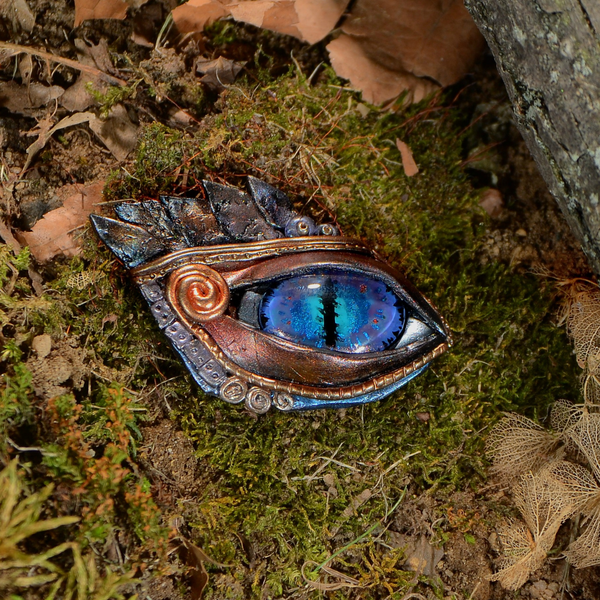 egyptian blue eye brooch with gold and bronze feather details and gold swirl at corner of the eye. on moss near a tree.
