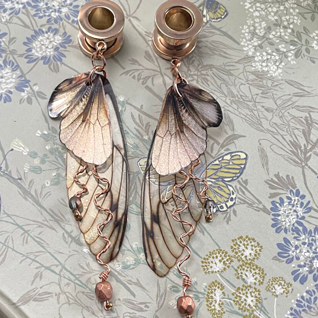 Double fairy wing earrings in brown with copper accents, with ear tunnels for stretched ears. Earrings are resting on a butterfly book