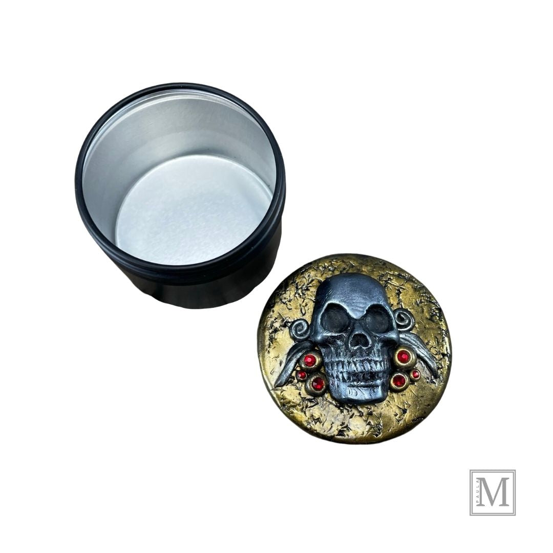 Lid covered with gold polymer clay with a silver skull, red crystals and polymer clay details in silver next to empty base container.