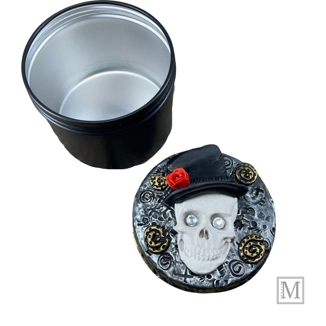 Polymer clay lid with a white skull with crystal eyes with top hat and red rose on a textured silver background with gold clay details.  Base of tin is next to decorative lid.