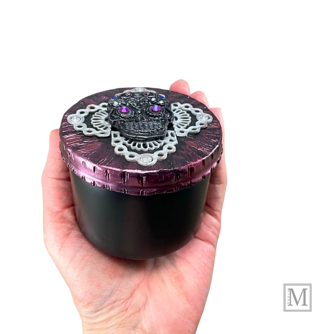 Black and Silver Sugar Skull with pink luster crystal eyes and colred small crystal details on a clay lace cross overlay with a deep metallic clay covered lid. Lid is propped up against black base of tin. Held in model's hand for size reference.