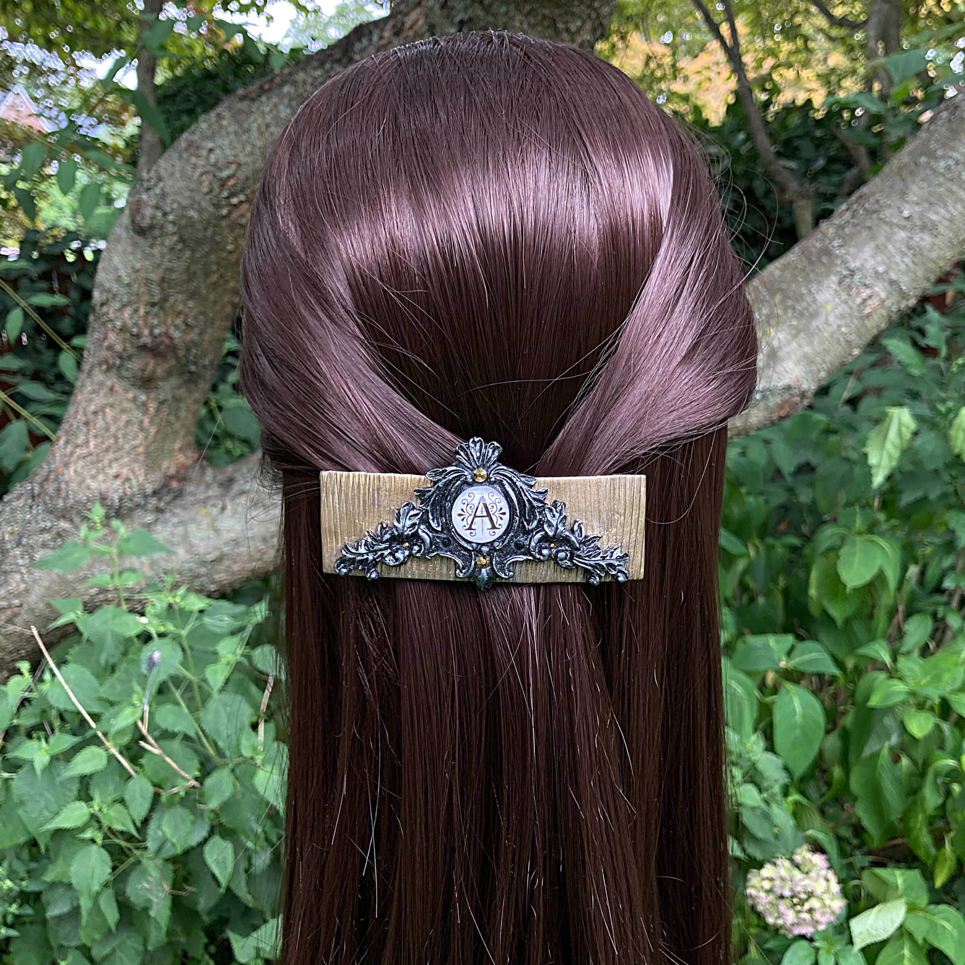 A model, in a garden,wearing a gold clay base hair clip with a filigree design and black and silver in the center a graphic with a letter A. 