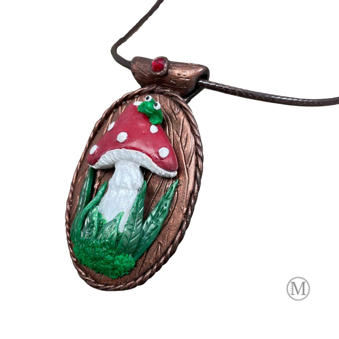 Oval pendant with red spotted mushroom with grass and moss at stem with little green frog sitting on top of the mushroom. Brown cord necklace through a tube bail with a red crystal