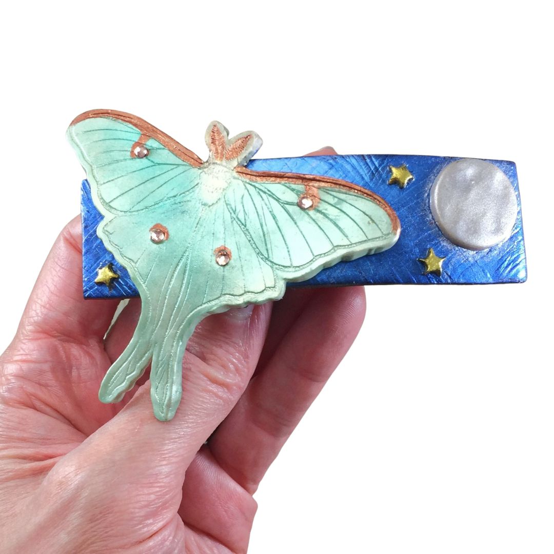 Polymer clay green giant silk moth on blue base with moon and stars hair clip held In hand.