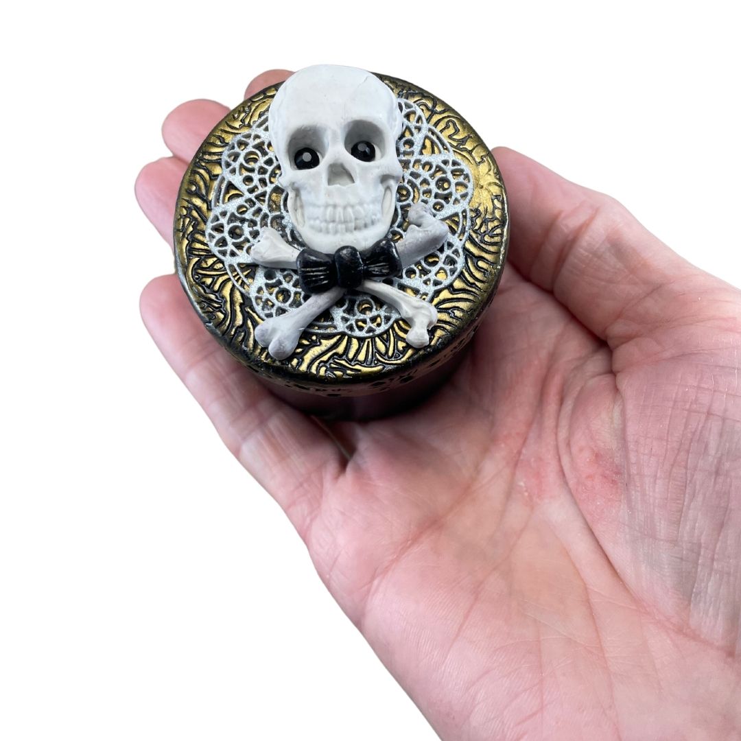 White skull and crossbones with black crystal eyes and dickie bow against white lace clay and gold textured lid of a storage jar in a hand for size reference