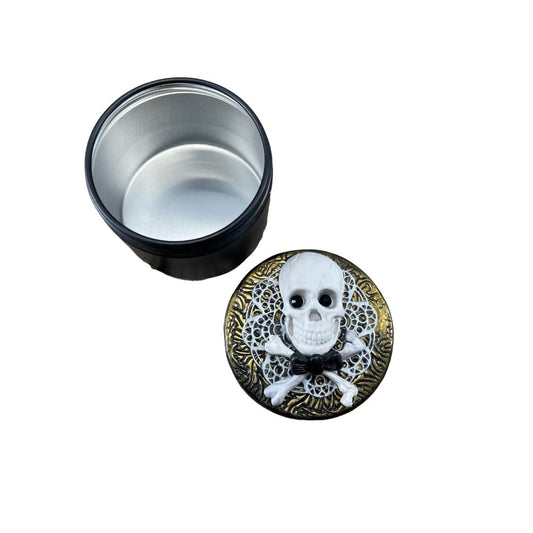 White skull and crossbones with black crystal eyes and dickie bow against white lace clay and gold textured lid of a storage jar. Lid is unscrewed and set to aside the storage tin.