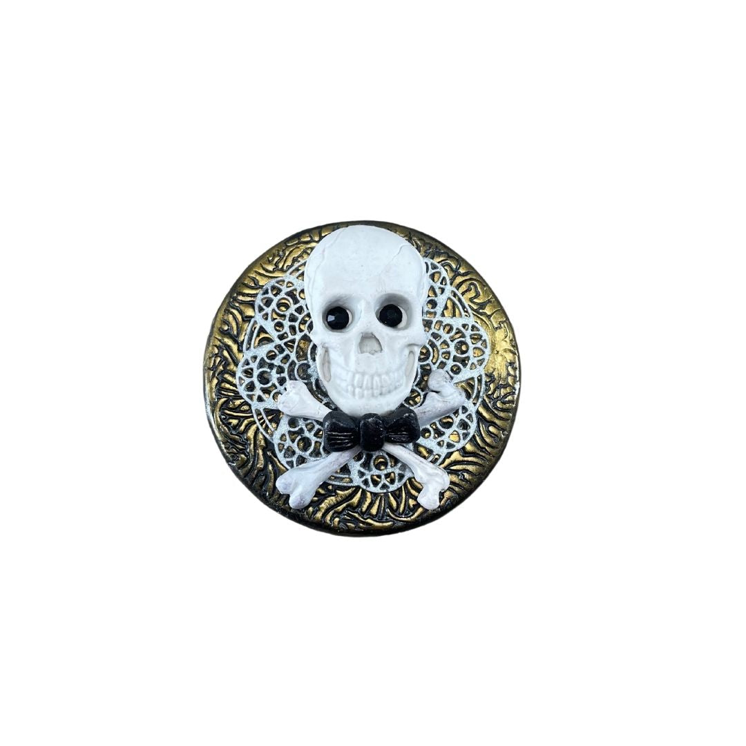 White skull and crossbones with black crystal eyes and dickie bow against white lace clay and gold textured lid of a storage jar.