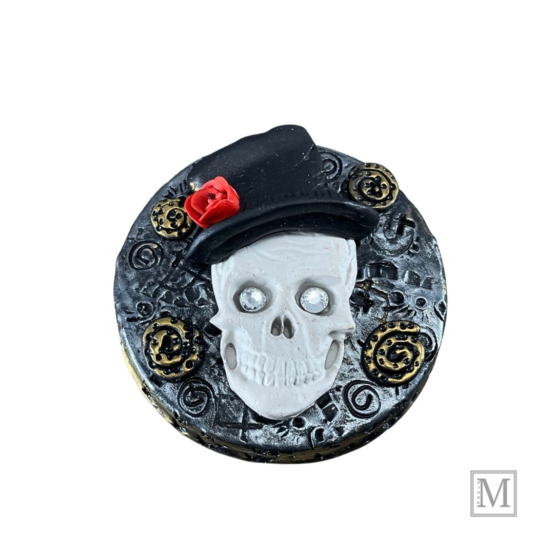 Polymer clay lid with a white skull with crystal eyes with top hat and red rose on a textured silver background with gold clay details. 