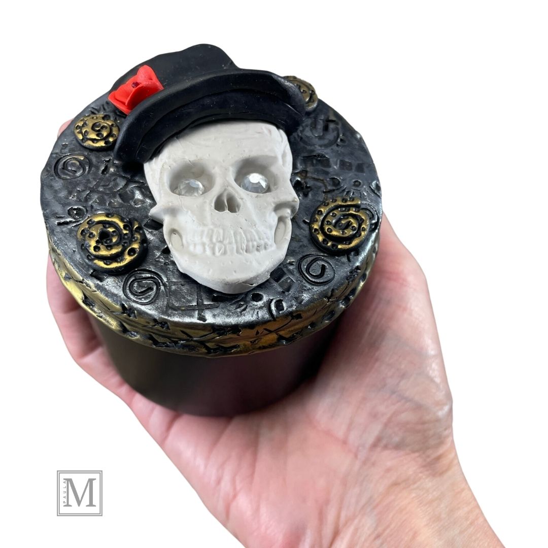 Polymer clay lid with a white skull with crystal eyes with top hat and red rose on a textured silver background with gold clay details. Tin is held in models hand.
