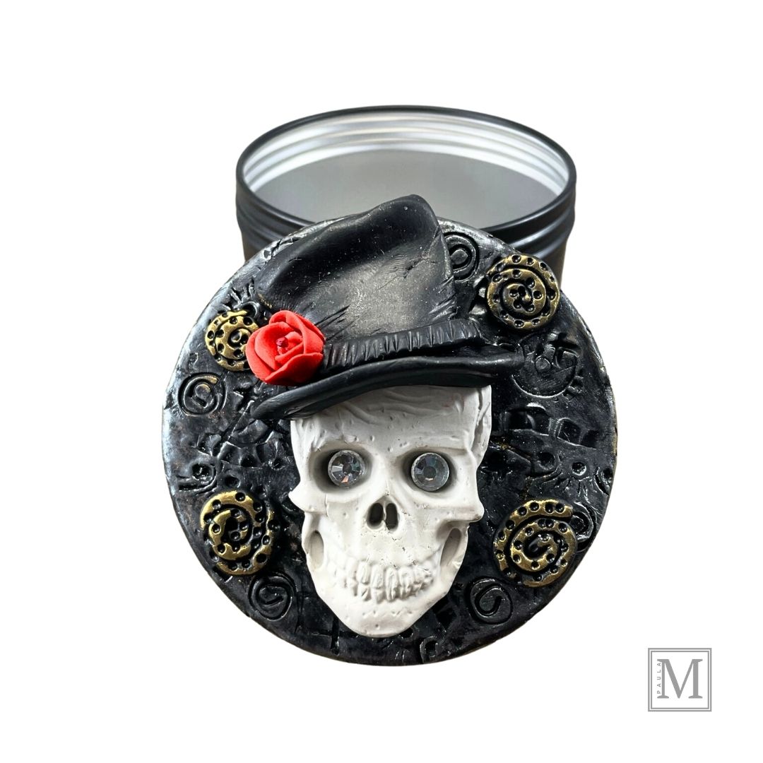 Polymer clay lid with a white skull with crystal eyes with top hat and red rose on a textured silver background with gold clay details. Base of tin is next to decorative lid.