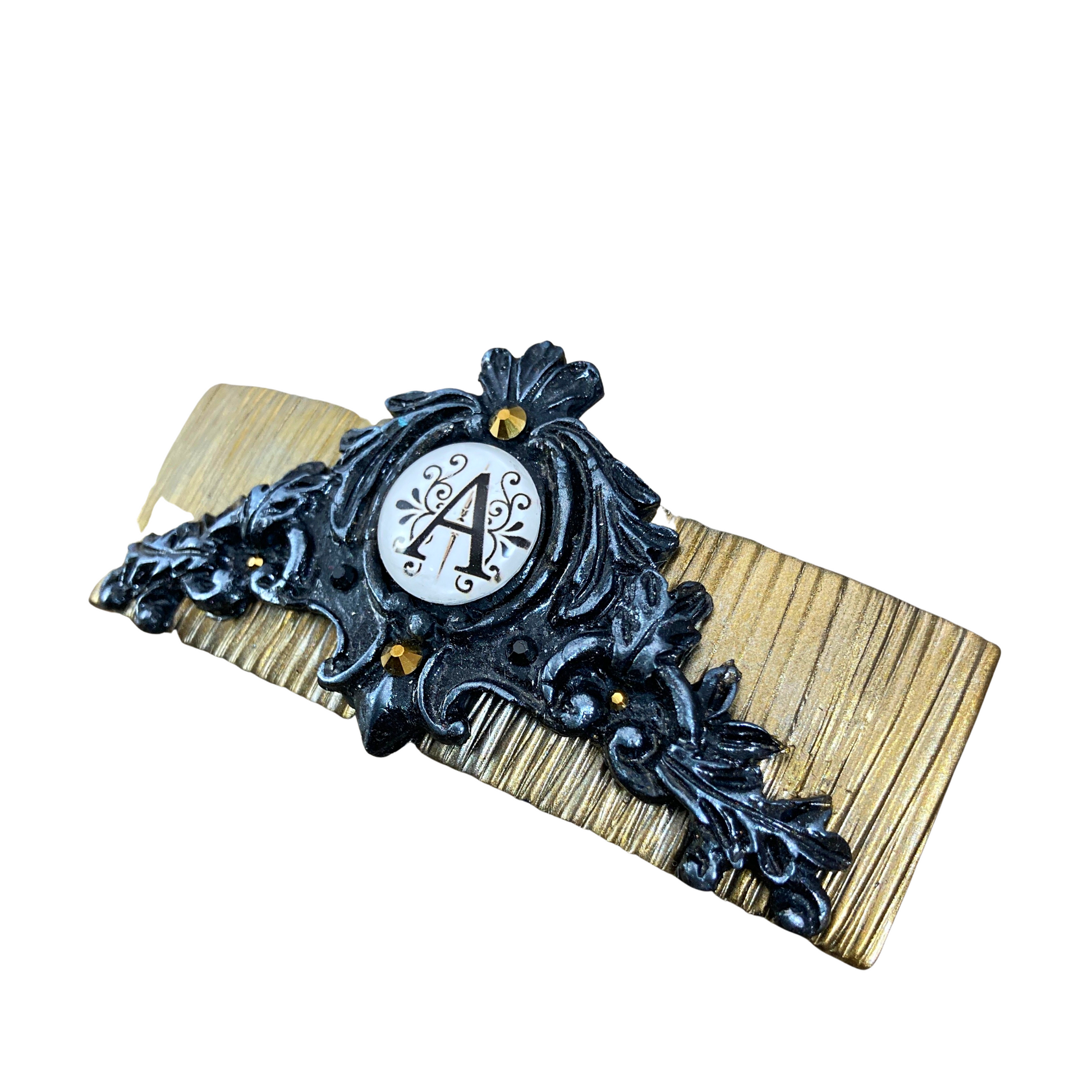 Side view of A gold clay base hair clip with a filigree design and black and silver in the center a graphic with a letter A.